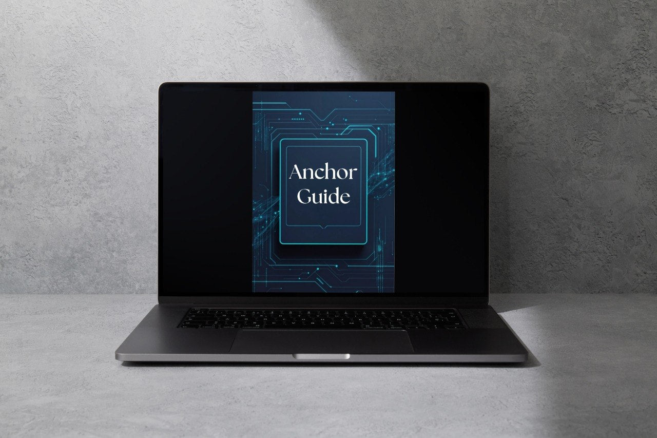 A dark gray laptop sits open on a light gray surface against a textured gray wall, with its screen displaying a blue graphic that reads 'Anchor Guide'.