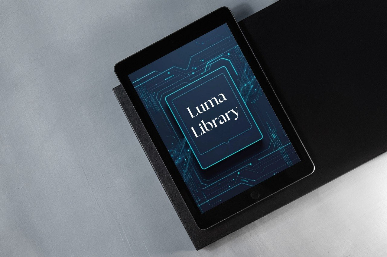 A black tablet device lying on a dark rectangular surface against a light gray background, with the screen displaying the words 'Luma Library' in white over a blue circuit-style graphic.